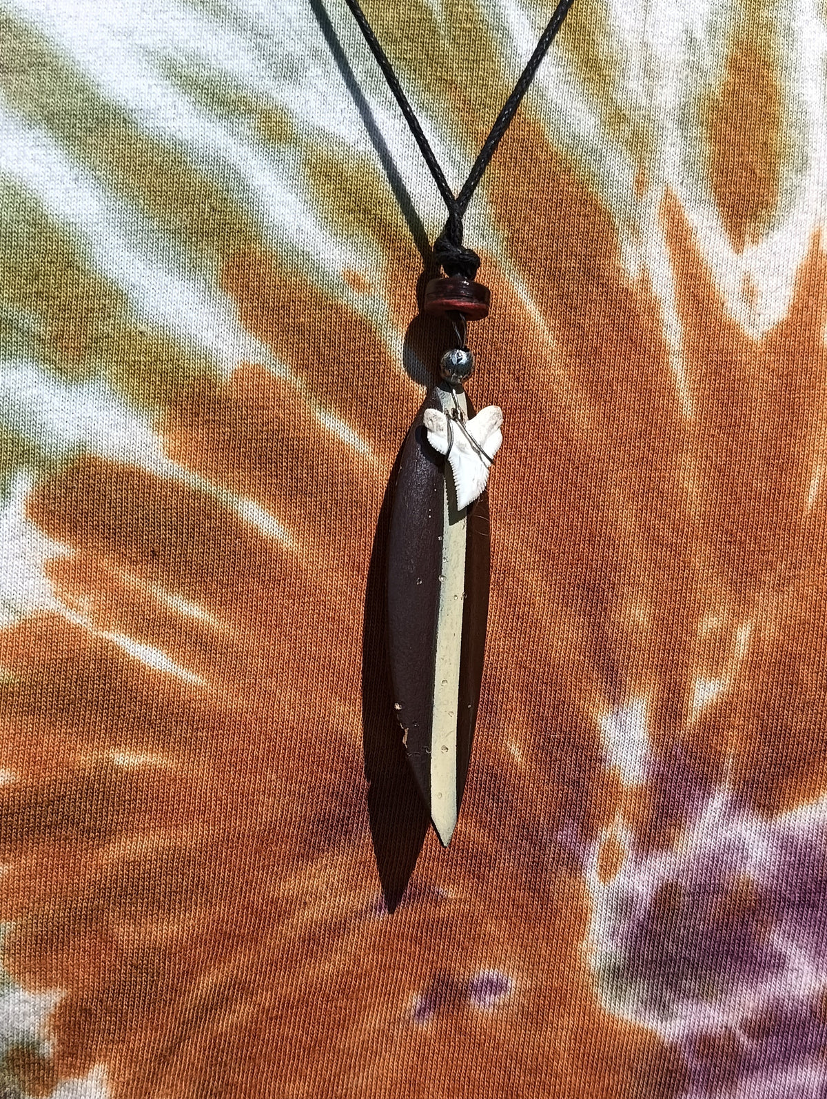 Shark Tooth and Wooden Surfboard Pendant Necklace – close-up showing natural shark tooth, dark wooden surfboard charm and waxed cord on tie-dye background